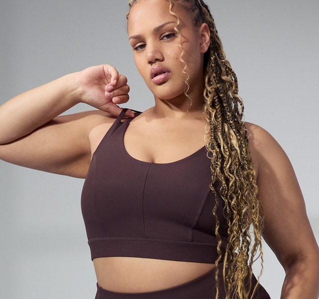 A woman with long golden-highlighted wavy braids poses against a soft gray background with blurred geometric window light, one arm raised with her fist near her chin. She wears a matching dark chocolate brown ribbed seamless athletic set consisting of a scoop-neck sports bra with a seamed center front detail and matching high-waist leggings visible at the bottom of the frame.