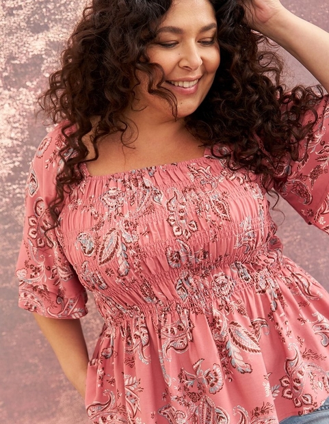 A close-up of a woman with voluminous dark curly hair smiling with her eyes cast downward, one hand raised to her head, against a textured rose-pink background. She wears a dusty rose smocked square-neck top with elbow-length flutter sleeves and a peplum hem, covered in a detailed white and gray paisley and floral print. The top features a fully smocked bodice from neckline to waist.