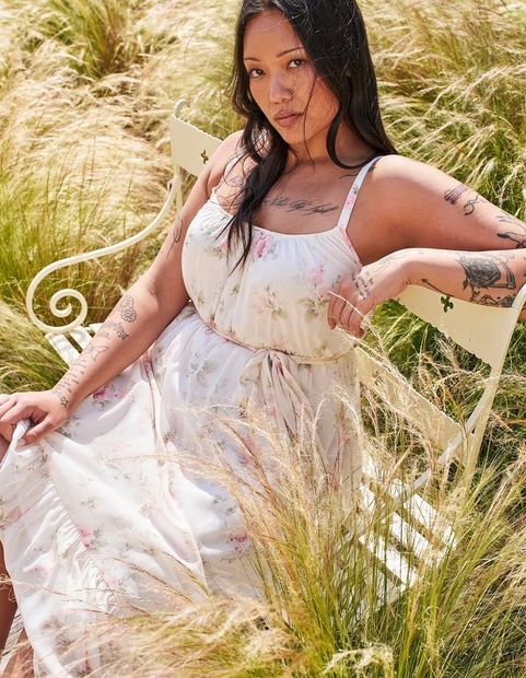 A woman with long straight dark hair sits on a white wrought iron garden chair surrounded by tall golden ornamental grass blowing in the breeze. She wears a cream spaghetti-strap maxi dress with a gathered scoop neckline, a drawstring waist tie, and a full skirt, printed with soft pink roses and green leaves. She holds the skirt out to one side. She has extensive tattoos on both arms and her chest, and looks directly at the camera with a calm expression.
