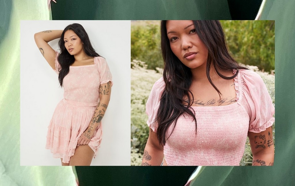 A wide horizontal banner with large green tropical leaves as the background. Two overlapping photo panels show the same woman with long straight dark hair and extensive tattoos on her arms, chest, and legs. In the left panel, she poses against a plain white background with one arm raised behind her head, wearing a light pink smocked square-neck mini dress with short puff sleeves, a fitted smocked bodice, and a tiered ruffle skirt. In the right panel, a close-up outdoor shot shows her from the chest up against a backdrop of white flowering plants and greenery, focusing on the detail of the smocked square neckline with a ruffled edge and short puff sleeves. A script chest tattoo is visible.