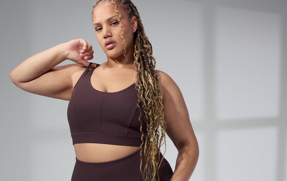 A woman with long golden-highlighted wavy braids poses against a soft gray background with blurred geometric window light, one arm raised with her fist near her chin. She wears a matching dark chocolate brown ribbed seamless athletic set consisting of a scoop-neck sports bra with a seamed center front detail and matching high-waist leggings visible at the bottom of the frame.