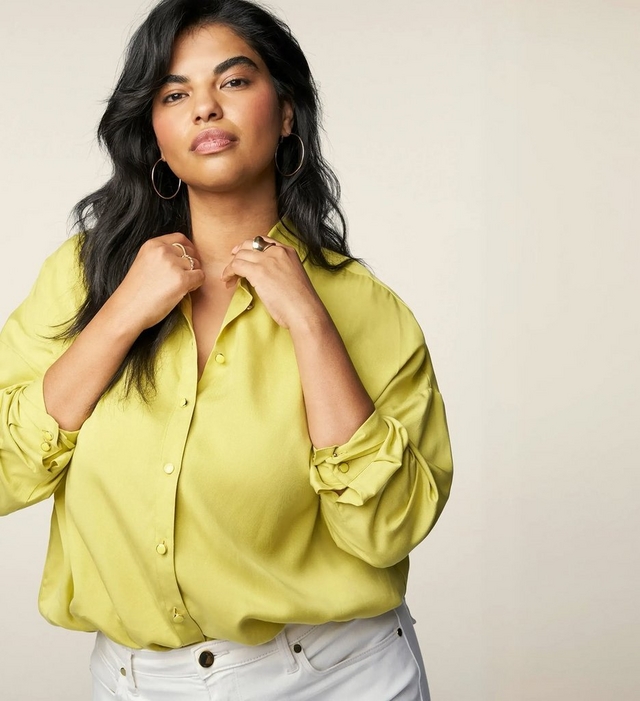 A close-up portrait of a woman with long dark wavy hair against a soft cream background, both hands raised to her collar. She wears a chartreuse yellow-green satin button-front shirt with the sleeves rolled up, paired with white jeans. She wears large gold hoop earrings and multiple rings on her fingers.