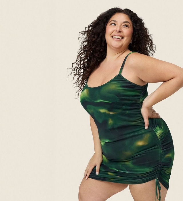 A woman with voluminous long dark curly hair laughs and looks upward against a soft cream background, one hand on her hip and the other resting on her thigh. She wears a dark green abstract swirl print satin mini dress with thin spaghetti straps, a scoop neckline, and ruched sides with an adjustable tie drawstring at the hem.