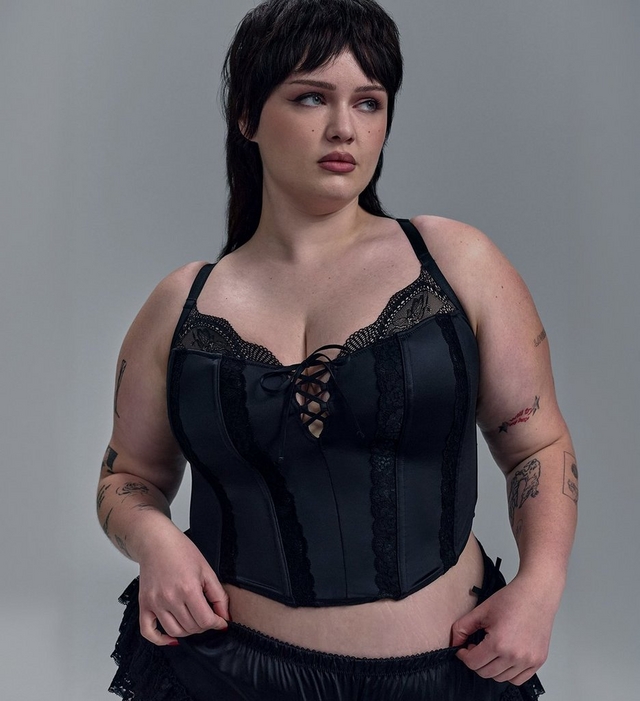 A closer crop of the same woman with short dark hair and wispy bangs looking to the side against a light gray background, both hands at her waist. She wears the same black satin corset-style bustier with boned panels, sheer black lace trim along the sweetheart neckline, a lace-up tie at the center front, lace trim along the seams, and thin shoulder straps. The top of a black satin and lace ruffle garment is visible at the bottom of the frame. Tattoos are visible on both arms.