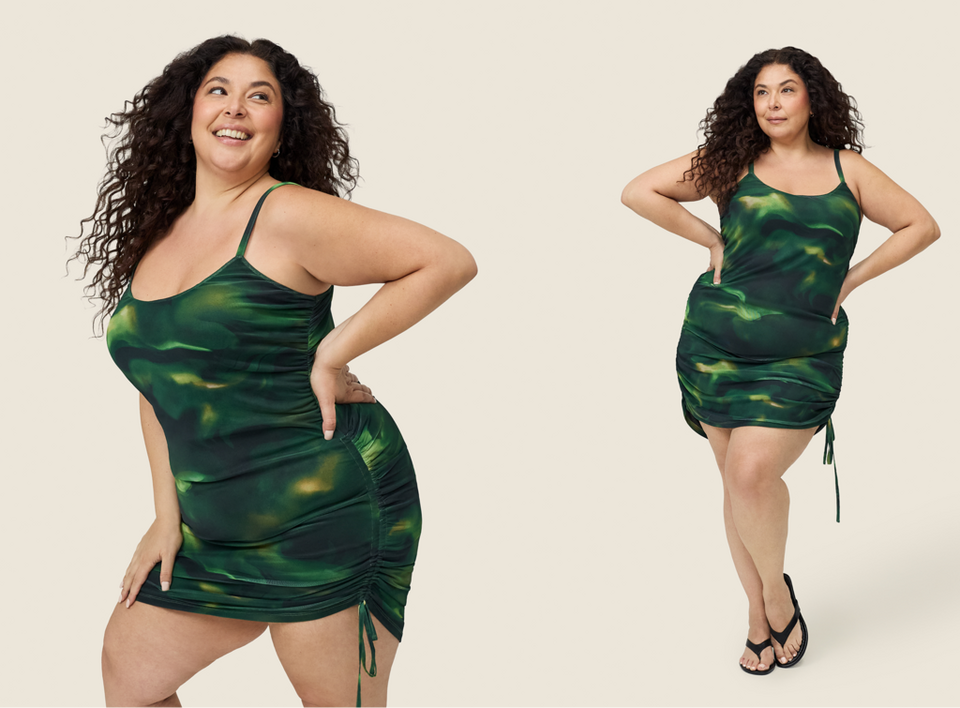 A wide horizontal banner against a soft cream background showing the same woman with voluminous long dark curly hair posed twice side by side. She wears a dark green abstract swirl print satin mini dress with thin spaghetti straps, a scoop neckline, ruched sides with adjustable tie drawstrings at the hem. In the left pose she laughs and looks upward with one hand on her hip. In the right pose she stands facing forward with both hands on her hips, wearing black thong flip flop sandals.