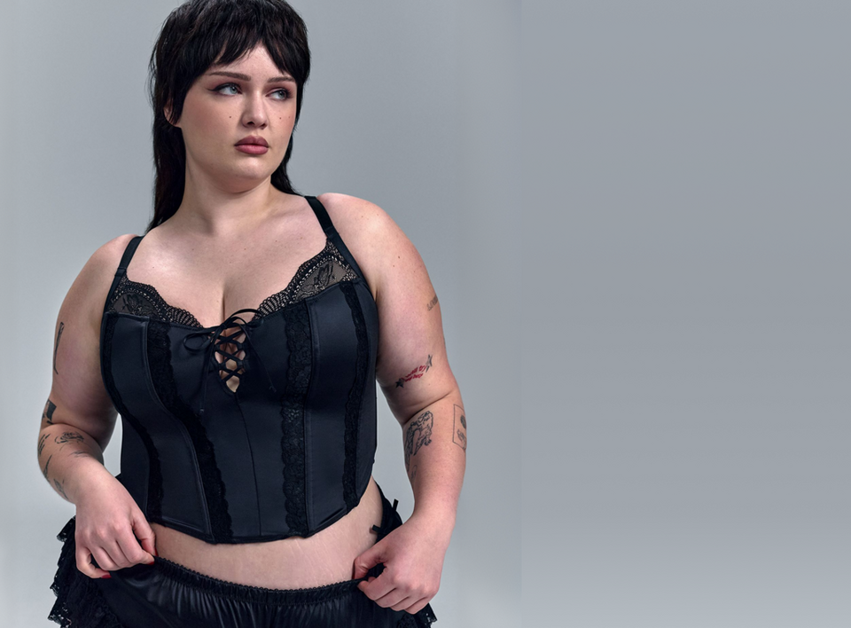 A woman with short dark hair and wispy bangs looks to the side against a light gray background, both hands at her waist. She wears a black satin corset-style bustier with boned vertical panels, sheer black lace trim along the sweetheart neckline, a lace-up tie detail at the center front, lace trim accents along the seams, and thin shoulder straps. The bottom of a black satin ruffle garment is partially visible at the hem. She has tattoos on both arms.