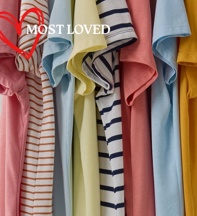 A close-up image of multiple t-shirts hanging tightly together, showing a colorful range of sleeve styles and fabrics. Visible colors and patterns from left to right include pink, white with brown horizontal stripes, light blue, pale yellow, white with navy blue stripes, coral pink, light blue, and mustard yellow. In the upper left corner, a red hand-drawn heart outline is overlaid with bold white text reading 'MOST LOVED TEES'.