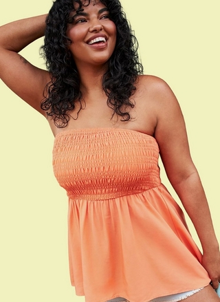 A woman with long dark curly hair smiles broadly while raising one hand behind her head. She wears a strapless smocked peach-orange tube top with a flowy peplum hem, paired with white frayed denim shorts partially visible at the bottom. The background is a solid pale yellow.