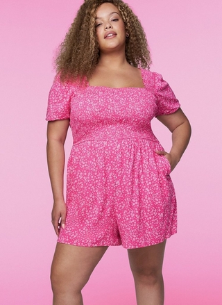 A woman with long curly light brown hair stands with one hand on her hip, looking slightly off camera. She wears a hot pink short romper with an allover small white floral print, a square neckline, smocked bodice, short puff sleeves, and a gathered skirt with visible side pockets. The background is a solid bright pink.