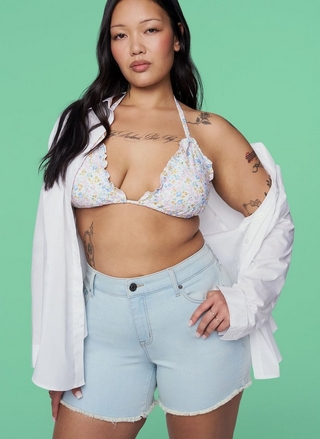 A woman with long straight dark hair gazes directly at the camera. She wears an open white button-down shirt layered over a white halter bikini top with a small floral print in pink, blue, and yellow and scalloped edges, paired with high-waisted light-wash denim cutoff shorts with a button closure and frayed hem. She has multiple tattoos on her chest, arms, and torso. She wears rings on her fingers. The background is a solid sage green.