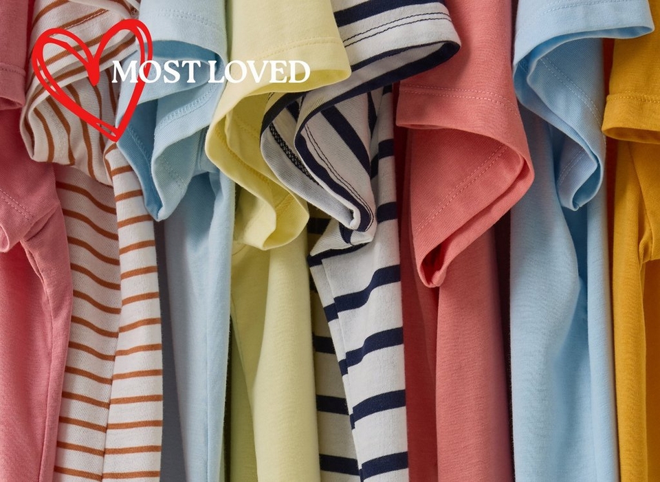 A close-up image of multiple t-shirts hanging tightly together, showing a colorful range of sleeve styles and fabrics. Visible colors and patterns from left to right include pink, white with brown horizontal stripes, light blue, pale yellow, white with navy blue stripes, coral pink, light blue, and mustard yellow. In the upper left corner, a red hand-drawn heart outline is overlaid with bold white text reading 'MOST LOVED TEES'.