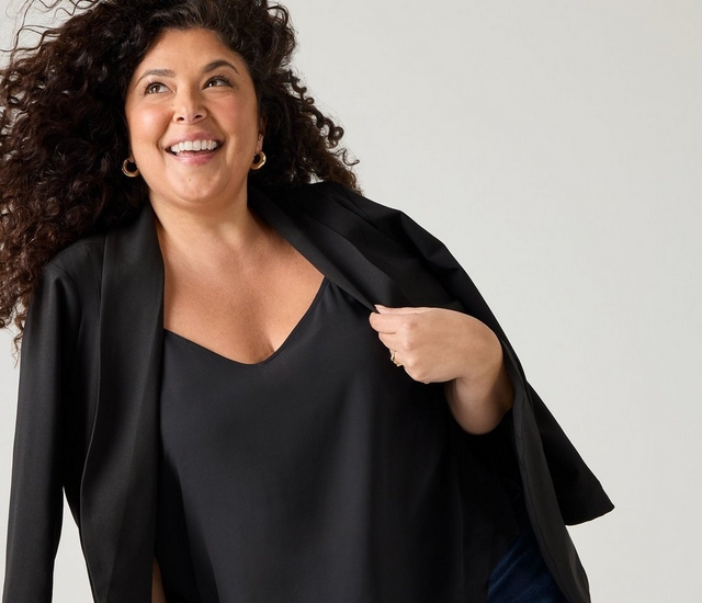 A woman with long curly dark brown hair smiles broadly, looking up and to the side, with her hair appearing windswept.A woman with long curly dark brown hair smiles broadly, looking up and to the side, with her hair appearing windswept. She wears gold hoop earrings, a gold ring, and a black V-neck top layered under an open black blazer that she holds open at the collar with one hand. A hint of dark denim is visible at the bottom of the image. The background is a plain light gray.