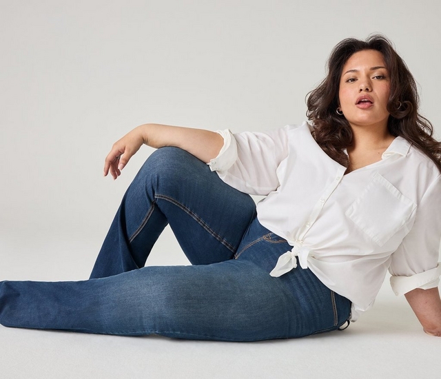 A woman with long wavy dark brown hair sits on the floor with one knee bent, leaning back on one arm, looking toward the camera. She wears small hoop earrings, an oversized white button-down shirt tied at the front, medium-wash skinny jeans, and black shoes. The background is a plain off-white.