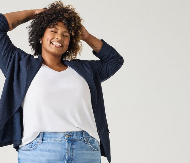 A woman with short curly dark hair with caramel highlights smiles with her eyes closed, both hands raised to her hair. She has a tattoo on one forearm, small hoop earrings, and wears a white V-neck t-shirt layered under an open navy pinstriped blazer, paired with light-wash denim jeans. The background is a plain off-white.