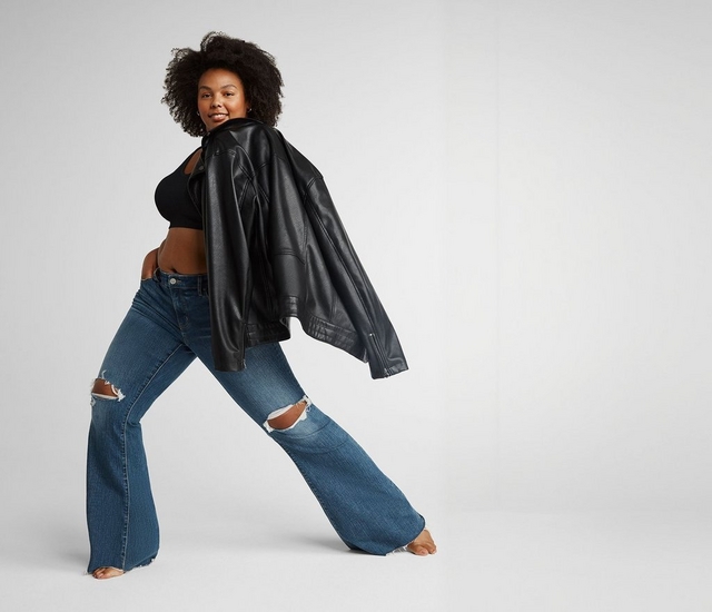 A woman with short curly dark hair stands in a wide lunging stance, looking back over her shoulder toward the camera with a slight smile. She wears small hoop earrings, a black cropped tank top, an oversized black faux leather jacket draped over her shoulders, and medium-wash flared jeans with distressed rip details at the knees. She is barefoot. The background is a plain off-white.