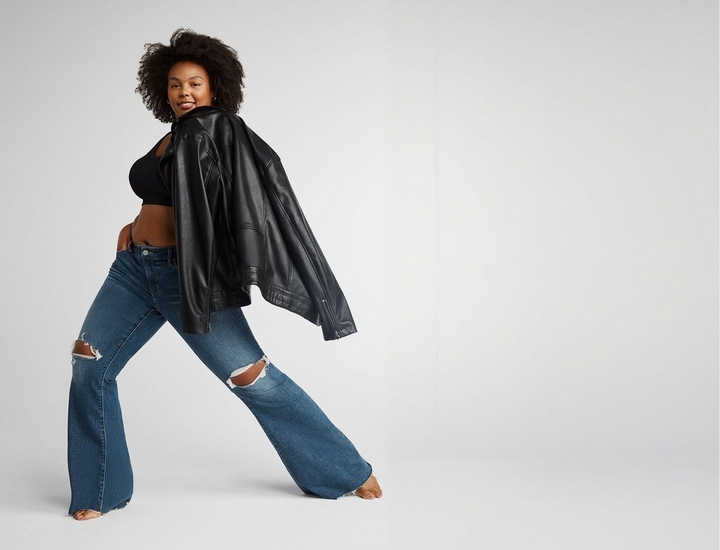 A woman with short curly dark hair stands in a wide lunging stance, looking back over her shoulder toward the camera with a slight smile. She wears small hoop earrings, a black cropped tank top, an oversized black faux leather jacket draped over her shoulders, and medium-wash flared jeans with distressed rip details at the knees. She is barefoot. The background is a plain off-white.