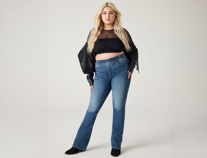 A woman with long wavy blonde hair stands with one hand on her hip, looking toward the camera. She wears a black mesh long-sleeve top with a ribbed black bandeau layered underneath, a black cropped faux leather jacket worn off the shoulders, medium-wash bootcut jeans, and black ankle boots. The background is a plain off-white.