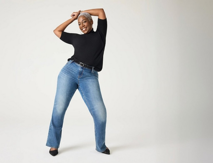 A woman with short curly gray hair smiles widely with her arms raised and crossed above her head in a playful dancing pose, one leg stepped forward.A woman with short curly gray hair smiles widely with her arms raised and crossed above her head in a playful dancing pose, one leg stepped forward. She wears gold hoop earrings, a black short-sleeve top, medium-wash bootcut jeans with a black belt featuring a silver buckle, and black pointed-toe heels. The background is a plain off-white.