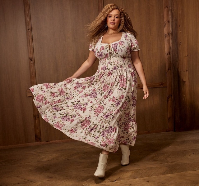 A woman with long curly light brown hair twirls in motion, one hand holding out the skirt of her dress, against a wood-paneled wall and wood floor background. She wears small hoop earrings and a long-sleeve midi dress with a sweetheart neckline trimmed in lace, a smocked waist, a flowing tiered skirt, all in a cream fabric with a pink and purple floral print and green leaves, paired with cream pointed-toe western-style ankle boots.
