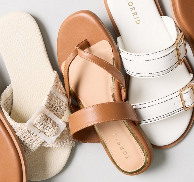 A flat-lay arrangement of several pairs of sandals against a light gray background, with a solid tan-colored circle in the upper left corner. The sandals include a tan thong sandal with a braided strap, a cream woven raglan-style slide sandal with a mother-of-pearl buckle, a tan leather thong sandal with a crossover strap and gold 'TORRID' branding on the insole, a white double-strap slide sandal with gold buckles and 'TORRID' branding, and a tan thong sandal with a braided strap and 'TORRID' embossed on the footbed.