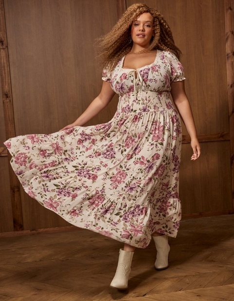 A woman with long curly light brown hair twirls in motion, one hand holding out the skirt of her dress, against a wood-paneled wall and wood floor background. She wears small hoop earrings and a long-sleeve midi dress with a sweetheart neckline trimmed in lace, a smocked waist, a flowing tiered skirt, all in a cream fabric with a pink and purple floral print and green leaves, paired with cream pointed-toe western-style ankle boots.
