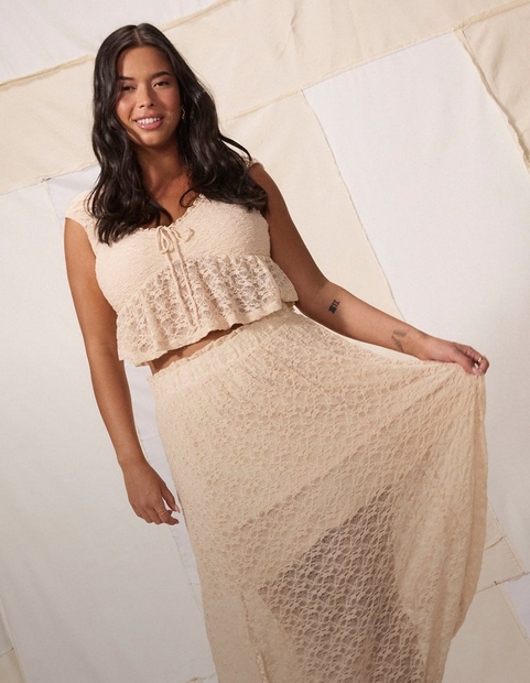 A woman with long dark wavy hair smiles at the camera while holding out the side of her skirt with one hand, against a cream and white patchwork fabric backdrop. She has a small tattoo on her forearm and wears a ring, small hoop earrings, and a cream lace two-piece set consisting of a cropped top with a tied neckline and ruffled hem, paired with a long sheer lace maxi skirt.