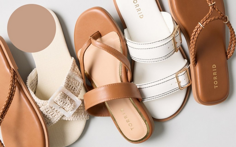 A flat-lay arrangement of several pairs of sandals against a light gray background, with a solid tan-colored circle in the upper left corner. The sandals include a tan thong sandal with a braided strap, a cream woven raglan-style slide sandal with a mother-of-pearl buckle, a tan leather thong sandal with a crossover strap and gold 'TORRID' branding on the insole, a white double-strap slide sandal with gold buckles and 'TORRID' branding, and a tan thong sandal with a braided strap and 'TORRID' embossed on the footbed.