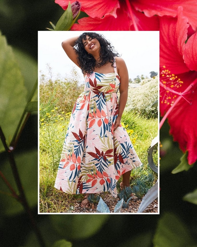A framed product photo with a white border set against a blurred background of red hibiscus flowers and green leaves. The photo shows the same woman from Image 1 standing outdoors in a garden, one hand raised to her hair, smiling with her eyes closed. She wears large gold hoop earrings and a sleeveless midi dress with a square neckline, fitted bodice, and full gathered skirt in a cream fabric with a large-scale tropical leaf print in red, orange, blue, olive green, and pink. She wears flat brown sandals and stands among large blue-gray agave plants, green grass, yellow wildflowers, and a pale sky in the background.