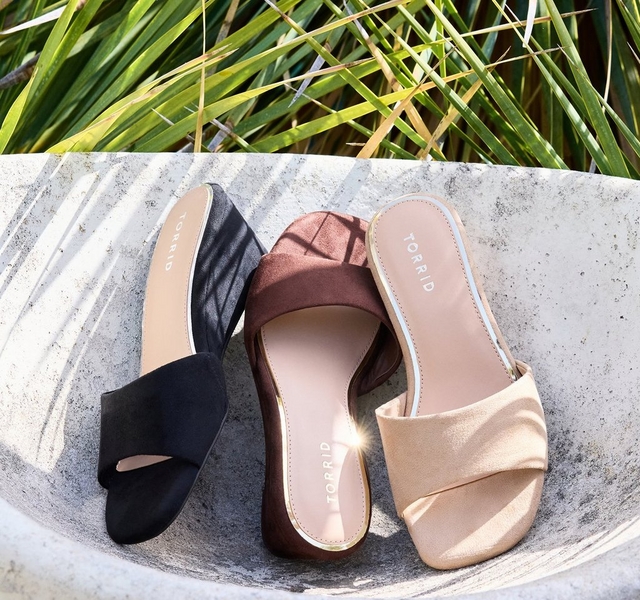 Three pairs of slide sandals arranged in a curved stone bowl with tropical grass blades visible in the background. From left to right: a black satin slide with a wide gathered toe strap and 'TORRID' branding on the insole, a dark brown suede slide with a wide gathered toe strap and 'TORRID' branding on the insole, and a champagne gold metallic slide with a wide gathered toe strap, white outsole, and 'TORRID' branding on the insole.