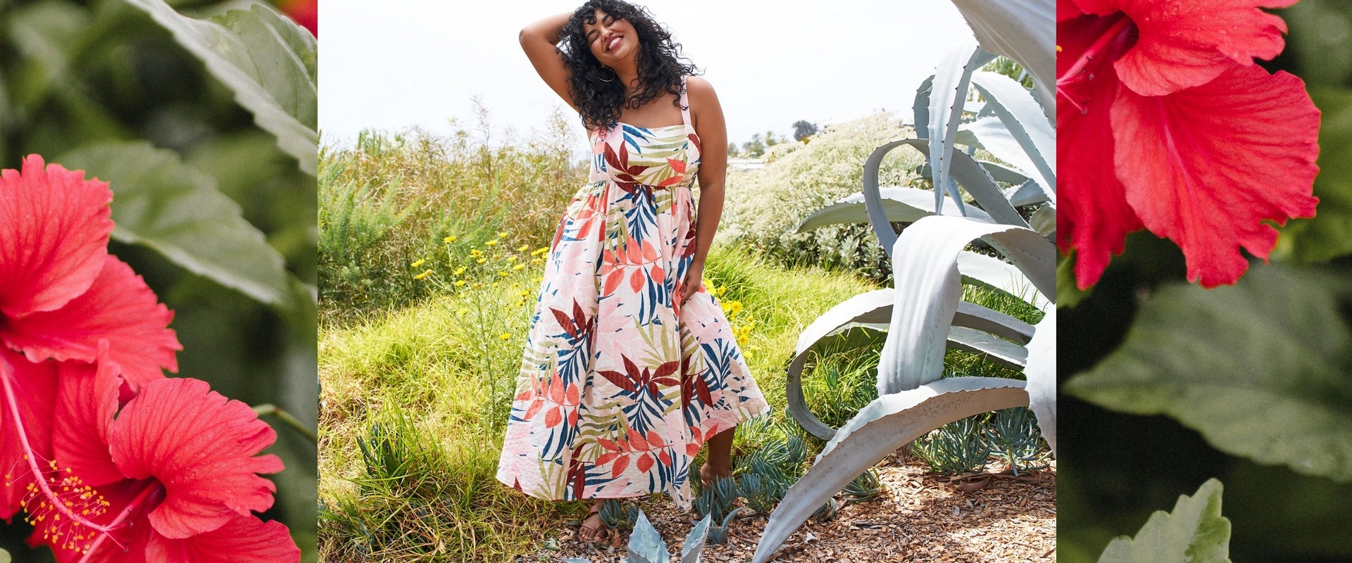 A three-panel banner image. The left and right panels show close-up photos of bright red hibiscus flowers with green leaves against a dark background. The center panel shows a woman with short curly dark hair standing outdoors in a garden setting, one hand raised to her hair, smiling with her eyes closed. She wears large gold hoop earrings and a sleeveless midi dress with a square neckline, fitted bodice, and full gathered skirt in a cream fabric with a large-scale tropical leaf print in red, orange, blue, olive green, and pink. She wears flat brown sandals. Behind her, large blue-gray agave plants, green grass, yellow wildflowers, and a pale sky are visible.