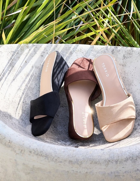 Three pairs of slide sandals arranged in a curved stone bowl with tropical grass blades visible in the background. From left to right: a black satin slide with a wide gathered toe strap and 'TORRID' branding on the insole, a dark brown suede slide with a wide gathered toe strap and 'TORRID' branding on the insole, and a champagne gold metallic slide with a wide gathered toe strap, white outsole, and 'TORRID' branding on the insole.