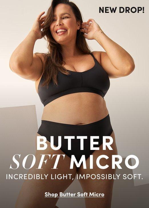A promotional banner with a warm light gray background. A woman with long dark wavy hair smiles with both hands raised to her hair. She wears a black seamless wireless bra with a scoop neckline and wide straps, and matching black high-waist bikini panties. Small black text in the upper right reads 'NEW DROP!' White and black text overlay at the bottom reads 'BUTTER SOFT MICRO' in large letters with smaller text reading 'INCREDIBLY LIGHT, IMPOSSIBLY SOFT.' and an underlined link reading 'Shop Butter Soft Micro.'
