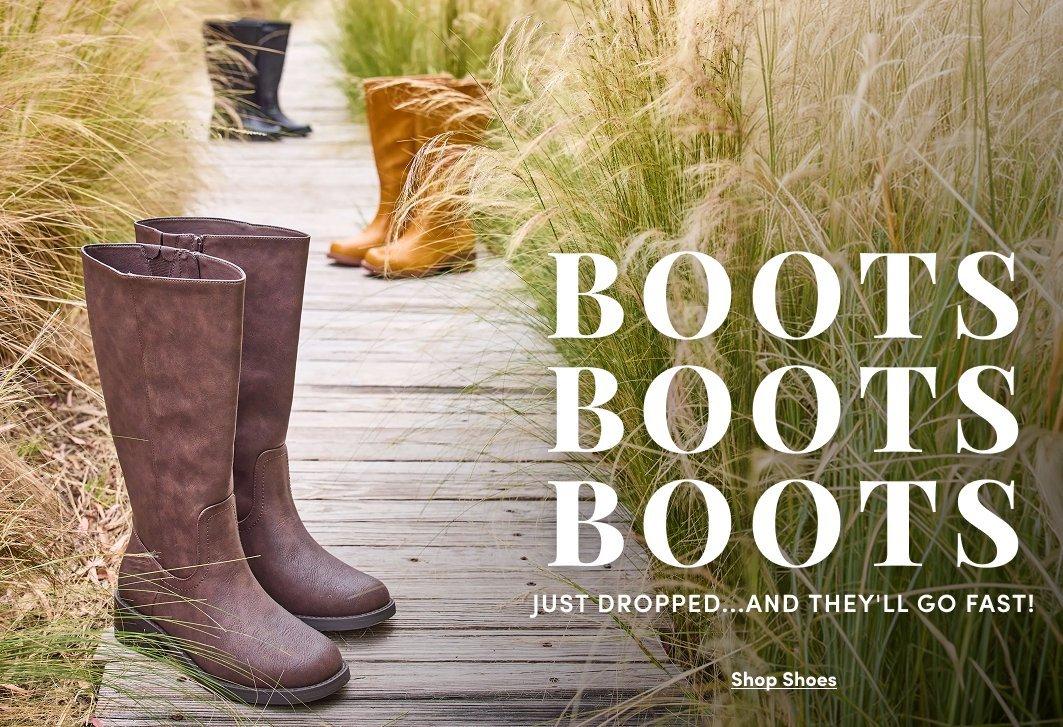 A promotional banner showing two pairs of tall riding-style boots standing on a weathered wooden dock or boardwalk, with tall dry grass and reeds visible in the background. One pair is dark brown and one is a lighter taupe brown, both with a classic tall shaft and low heel. Large bold black text to the right reads 'BOOTS BOOTS BOOTS' with smaller text below reading 'JUST DROPPED...AND THEY'LL GO FAST!' and a small link reading 'Shop Shoes.'