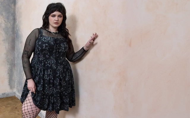 A model with long auburn hair against a light grey background wearing a black lace-up corset-style mini dress with grommet hardware and a layered black tulle ruffled skirt, holding the skirt out to the side.