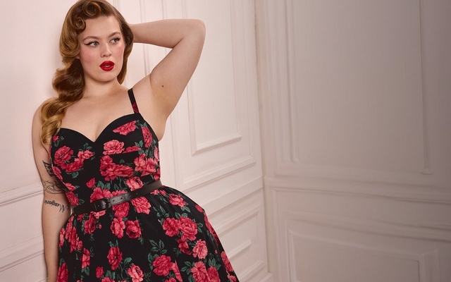 A model with pin-up style wavy brown hair and red lipstick poses against a white paneled wall wearing a blue fit-and-flare dress with a hot air balloon print, sweetheart neckline, wide straps, and a full pleated skirt.