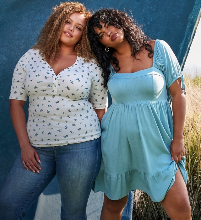 Two women stand together leaning against a dark teal painted backdrop with tall dry grass visible to the right. The woman on the left has long curly light brown hair, wears a ring, and is dressed in a cream ribbed henley top with a small blue ditsy floral print and elbow-length sleeves, paired with medium-wash straight leg jeans. The woman on the right has short curly dark hair with curtain bangs, wears gold hoop earrings and a ring, and is dressed in a solid light teal short dress with a square neckline, short puff sleeves, a fitted bodice, and a tiered ruffle hem.