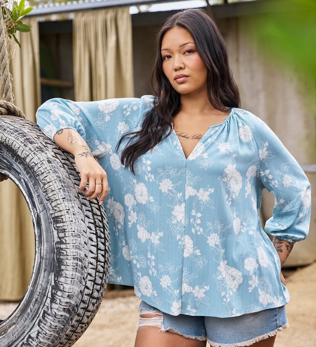 Two women stand together leaning against a dark teal painted backdrop with tall dry grass visible to the right. The woman on the left has long curly light brown hair, wears a ring, and is dressed in a cream ribbed henley top with a small blue ditsy floral print and elbow-length sleeves, paired with medium-wash straight leg jeans. The woman on the right has short curly dark hair with curtain bangs, wears gold hoop earrings and a ring, and is dressed in a solid light teal short dress with a square neckline, short puff sleeves, a fitted bodice, and a tiered ruffle hem.