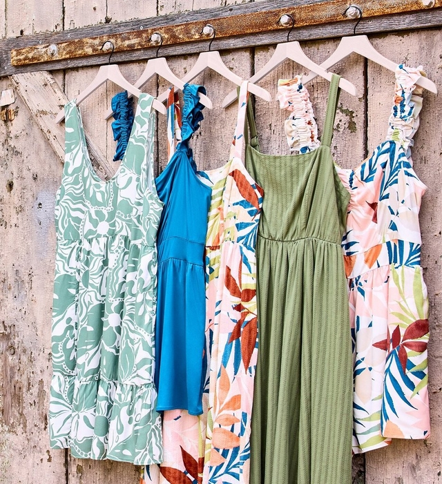 Five dresses hanging on blush pink velvet hangers on a rustic weathered wood barn door. From left to right: a sage green and white abstract floral tiered midi dress with ruffle shoulder straps; a solid teal blue tiered midi dress with ruffle halter straps; a cream midi dress with a large-scale tropical leaf print in orange, red, teal, green, and pink with ruched shoulder straps; a solid olive green maxi dress with a textured ribbed fabric, square neckline, and elastic waist; and a white midi dress with a large-scale tropical leaf print in blue, green, orange, and red with ruched shoulder straps.