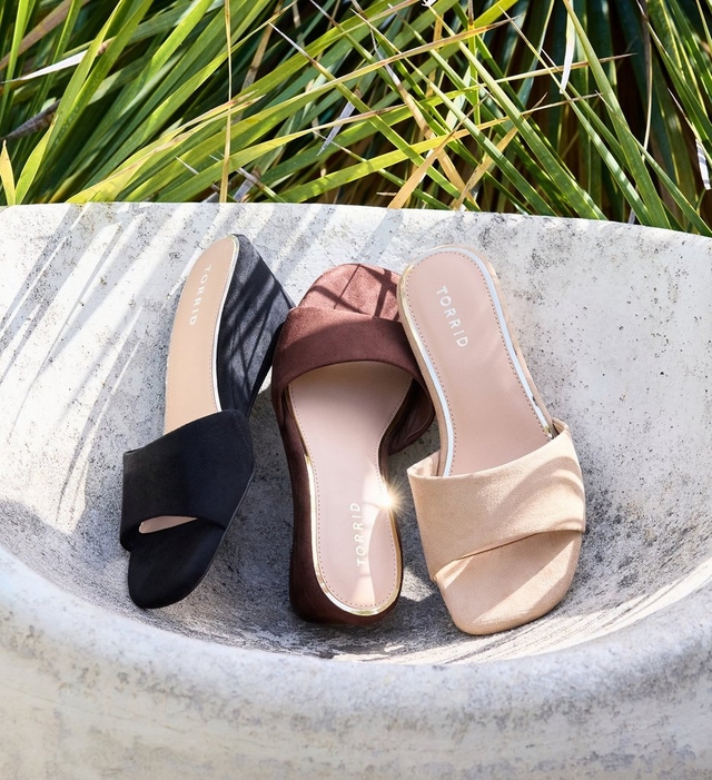 Three pairs of flat slide sandals arranged in a curved stone bowl with tropical grass blades visible in the background. From left to right: a black satin slide with a wide gathered toe strap and 'TORRID' branding on the insole; a dark brown suede slide with a wide gathered toe strap and 'TORRID' branding on the insole; and a champagne gold metallic slide with a wide gathered toe strap, white outsole, and 'TORRID' branding on the insole.