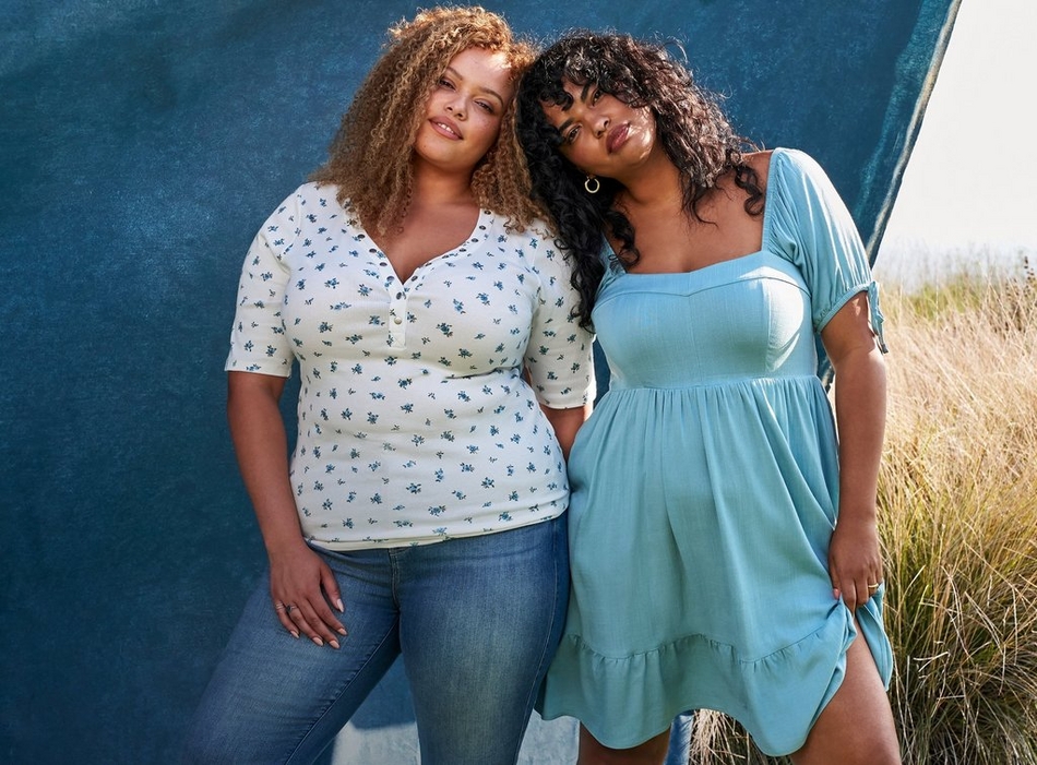 Two women stand together leaning against a dark teal painted backdrop with tall dry grass visible to the right. The woman on the left has long curly light brown hair, wears a ring, and is dressed in a cream ribbed henley top with a small blue ditsy floral print and elbow-length sleeves, paired with medium-wash straight leg jeans. The woman on the right has short curly dark hair with curtain bangs, wears gold hoop earrings and a ring, and is dressed in a solid light teal short dress with a square neckline, short puff sleeves, a fitted bodice, and a tiered ruffle hem.