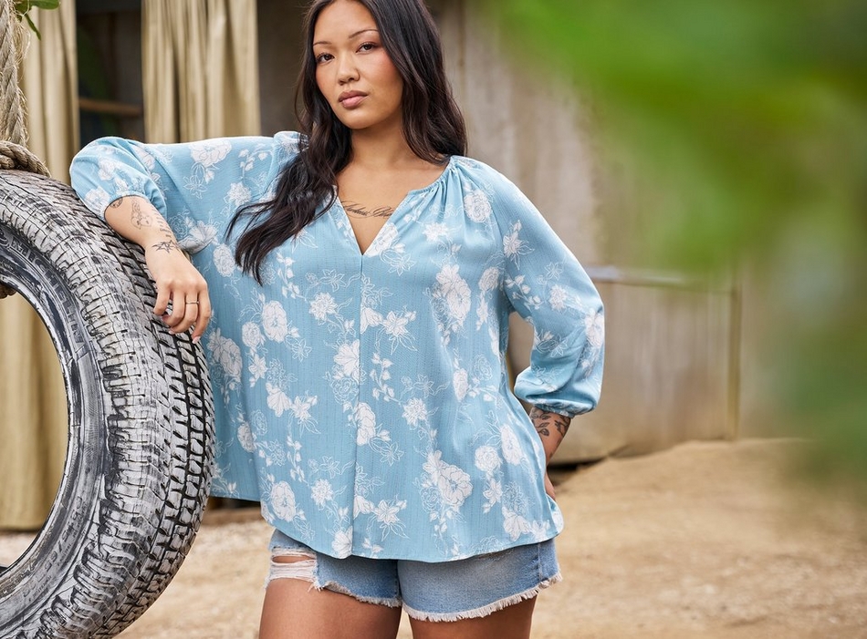 A woman with long straight dark hair leans against a large tire swing with one hand on her hip, looking toward the camera with a neutral expression. She has a script chest tattoo and tattoos on both forearms, and wears a ring. She is dressed in a light blue three-quarter sleeve blouse with a gathered V-neckline, voluminous puffed sleeves, and an allover white rose and floral print, paired with light-wash distressed denim cutoff shorts with frayed hems. A sandy outdoor area with a wood structure and green foliage is visible in the background.