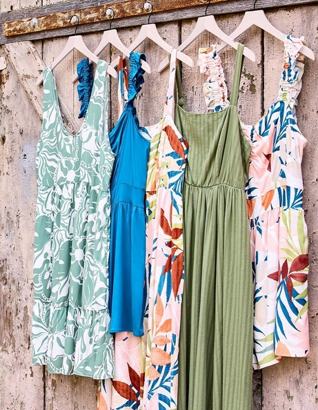 Five dresses hanging on blush pink velvet hangers on a rustic weathered wood barn door. From left to right: a sage green and white abstract floral tiered midi dress with ruffle shoulder straps; a solid teal blue tiered midi dress with ruffle halter straps; a cream midi dress with a large-scale tropical leaf print in orange, red, teal, green, and pink with ruched shoulder straps; a solid olive green maxi dress with a textured ribbed fabric, square neckline, and elastic waist; and a white midi dress with a large-scale tropical leaf print in blue, green, orange, and red with ruched shoulder straps.