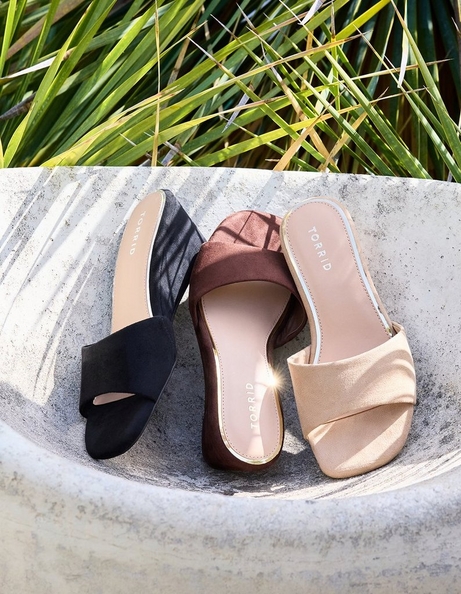 Three pairs of flat slide sandals arranged in a curved stone bowl with tropical grass blades visible in the background. From left to right: a black satin slide with a wide gathered toe strap and 'TORRID' branding on the insole; a dark brown suede slide with a wide gathered toe strap and 'TORRID' branding on the insole; and a champagne gold metallic slide with a wide gathered toe strap, white outsole, and 'TORRID' branding on the insole.