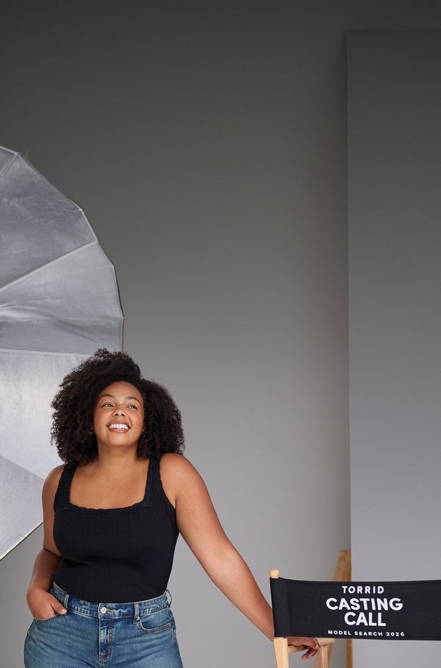 A woman with short natural curly dark hair stands smiling with one hand in her pocket next to a black director's chair labeled 'TORRID CASTING CALL MODEL SEARCH 2025' in white text. She wears a black square-neck tank top and light-wash denim jeans. A large silver photography umbrella reflector is visible to her left. The background is a plain gray studio setting.