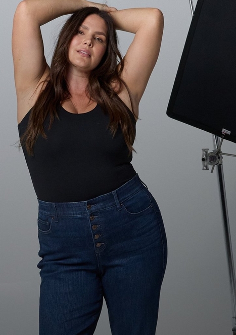 A woman with long straight dark brown hair stands with both arms raised above her head, looking toward the camera. She wears a black sleeveless square-neck top and dark wash denim jeans with a row of black exposed buttons at the waist. A black photography light panel on a stand is visible to her right. The background is a plain light gray.