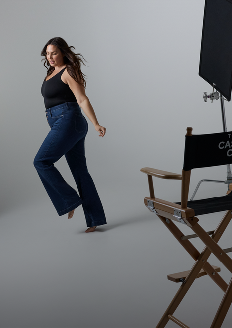 A woman with long straight dark brown hair appears to be mid-stride, looking down with her hair in motion. She is barefoot and wears a black sleeveless square-neck top and dark wash high-waist bootcut jeans. To her right, a tall wooden director's chair with a black canvas seat and back panel is partially visible, with text on the back partially reading 'CAS...' and 'C...' A black photography light panel on a metal stand is visible in the upper right corner. The background is a plain light gray studio floor and wall.