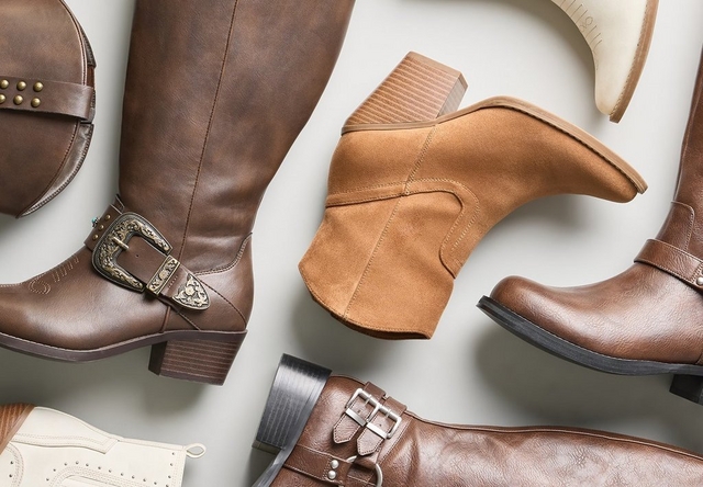 Close-up flat lay style photo of an assortment of brown and cream colored boots and booties arranged on a light gray background. Included are a tan suede pointed toe bootie with a stacked heel, a brown leather knee high boot with a buckled ankle strap and harness ring, another brown leather tall boot with an ornate engraved buckle strap, a cream colored perforated bootie, and a dark brown leather boot with a decorative buckle in the foreground.