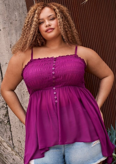 Photo of a woman with curly reddish blonde hair and freckles standing against a rustic wooden wall and a corrugated rust colored metal panel. She wears a magenta pink smocked cami top with thin straps, a ruffled trim neckline, a row of buttons down the front, and a flowy asymmetrical hem, paired with light wash denim shorts. Her hands rest on her hips.
