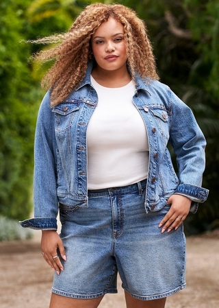 Photo of a woman with long curly reddish blonde hair standing outdoors with green foliage in the background. She wears a light wash denim jacket with studded details on the chest pockets and cuffed sleeves, open over a white ribbed tank top, paired with light wash denim shorts. She has rings on both hands and rests one hand on her hip.