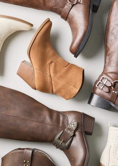 Close-up flat lay style photo of an assortment of brown and cream colored boots and booties arranged on a light gray background. Included are a tan suede pointed toe bootie with a stacked heel, a brown leather knee high boot with a buckled ankle strap and harness ring, another brown leather tall boot with an ornate engraved buckle strap, a cream colored perforated bootie, and a dark brown leather boot with a decorative buckle in the foreground.